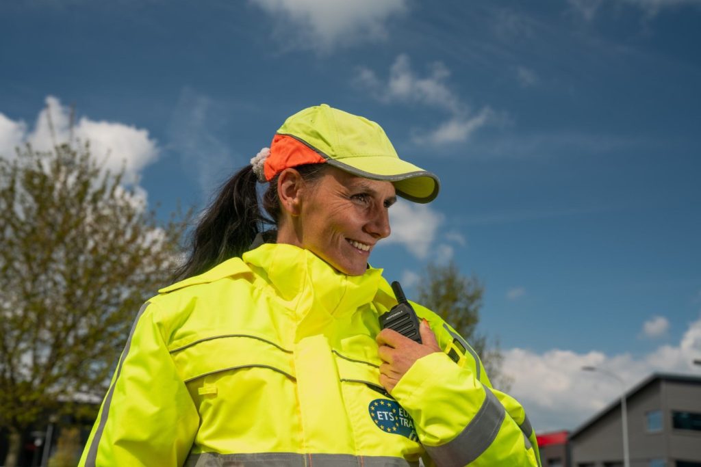 Vrouwelijke verkeersregelaar in felgele veiligheidskleding met portofoon, glimlachend onder een blauwe lucht.